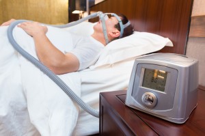 CPAP machine sitting on a bedside table with blurred Middle age asian man sleeping in his bed wearing CPAP mask in the background